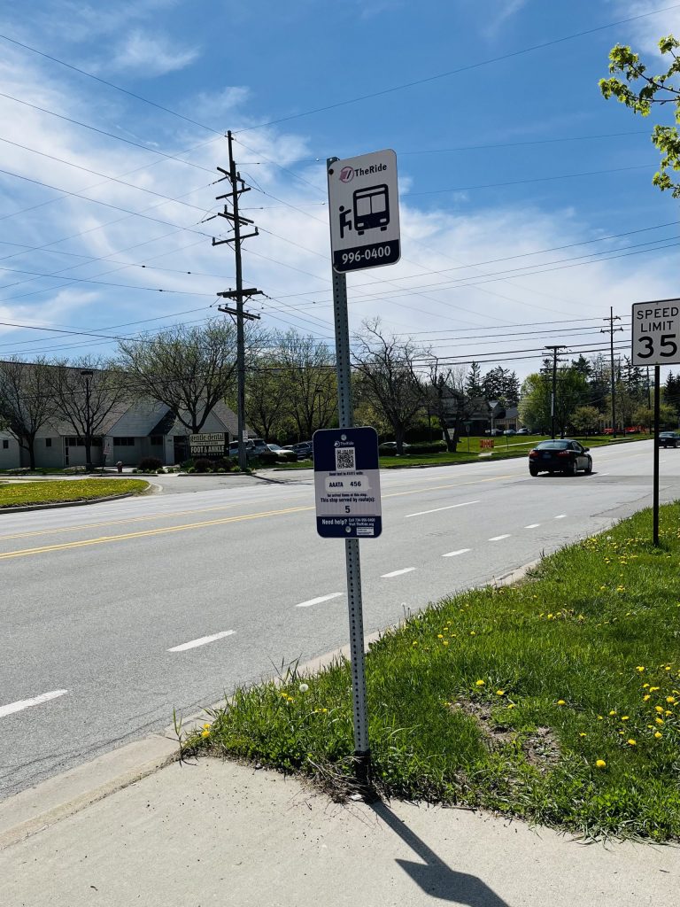 TheRide bus stop sign for route 5 on a grassy strip beside Packard Road.